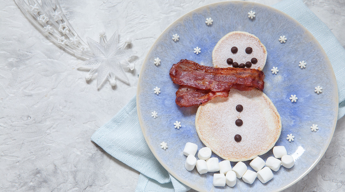 Snowman shaped pancake with chocolate chips, a bacon scarf, and marshmallow snow and snowflakes on a plate.